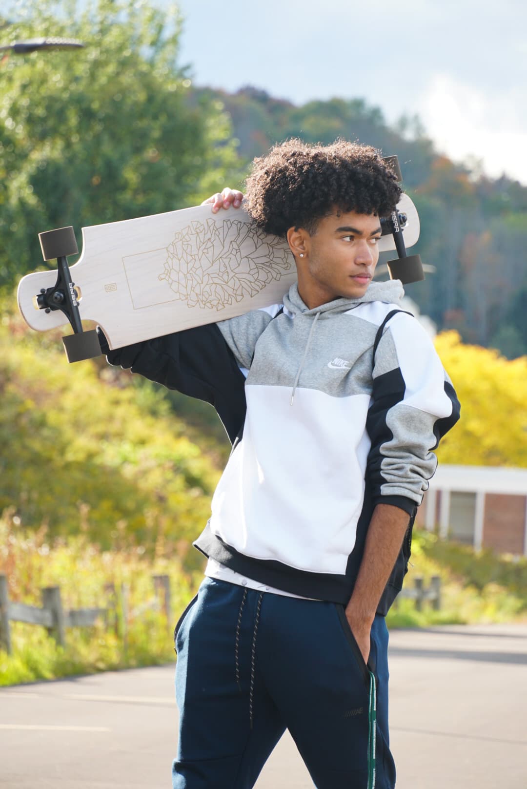 Solo portrait of Xavier carrying a longboard on his shoulder with fall foliage behind him